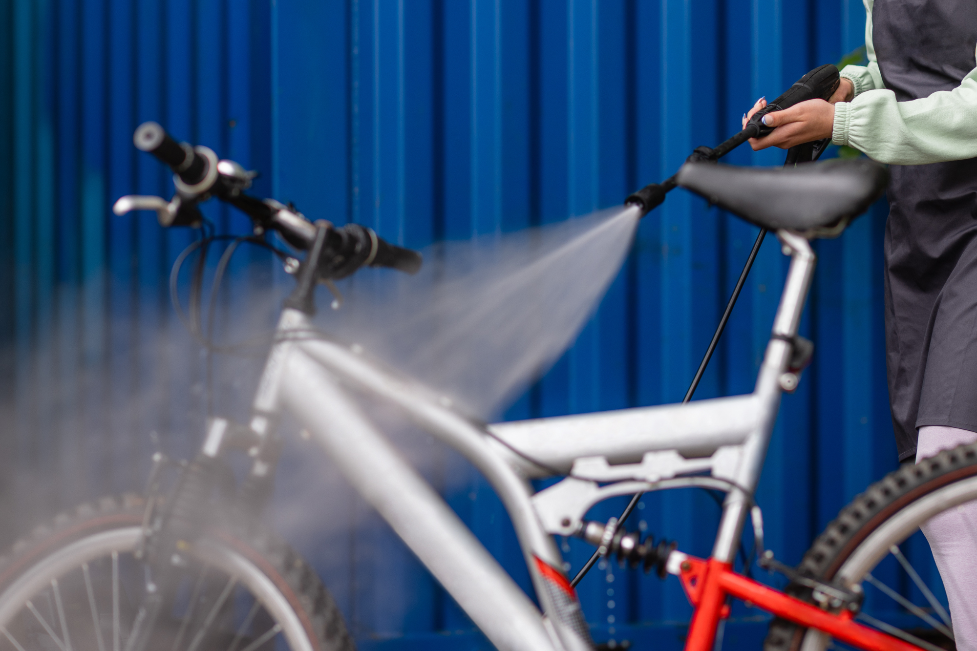 Person washing a bicycle with a hose against a blue corrugated metal background