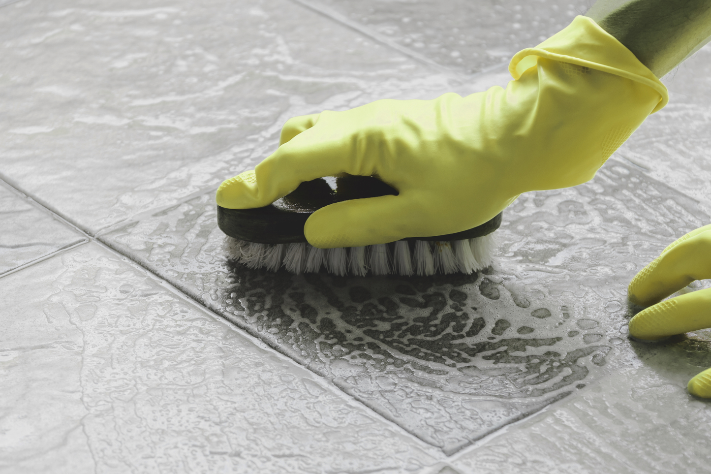 Person cleaning a tile floor with a brush and yellow gloves