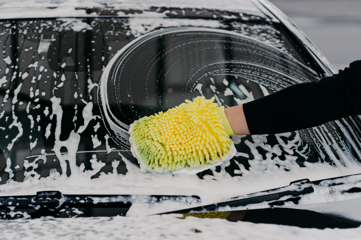 A close-up of a hand using a yellow and green microfiber cleaning sponge to wash a soapy black car window.