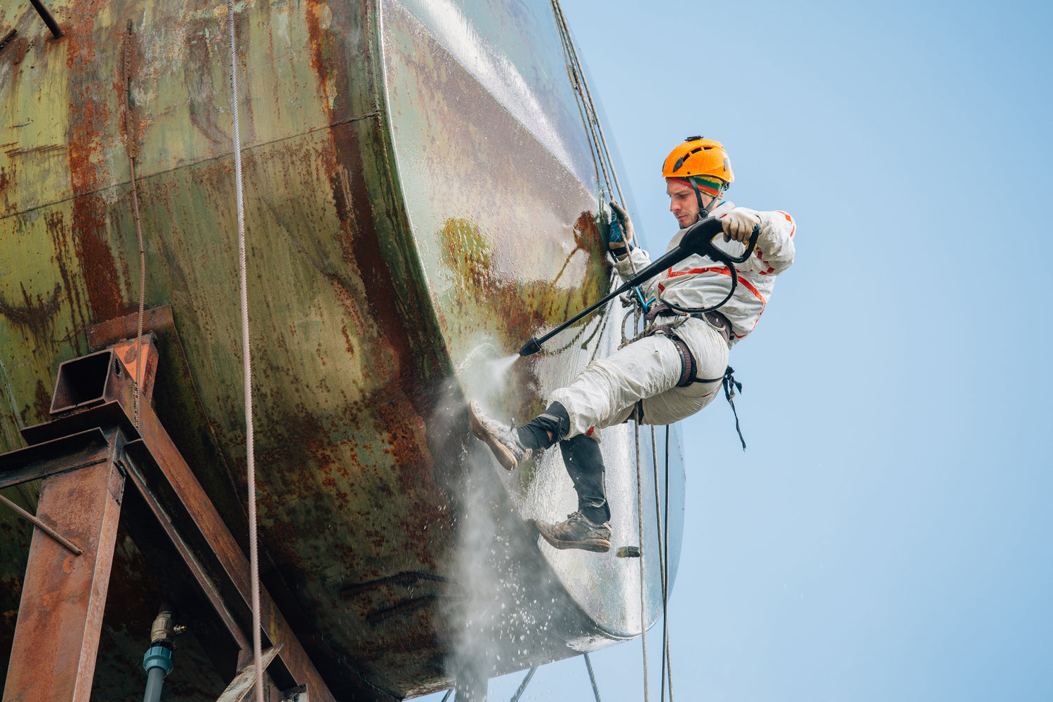 Person in safety gear cleaning a large metal tank with a blue sky background