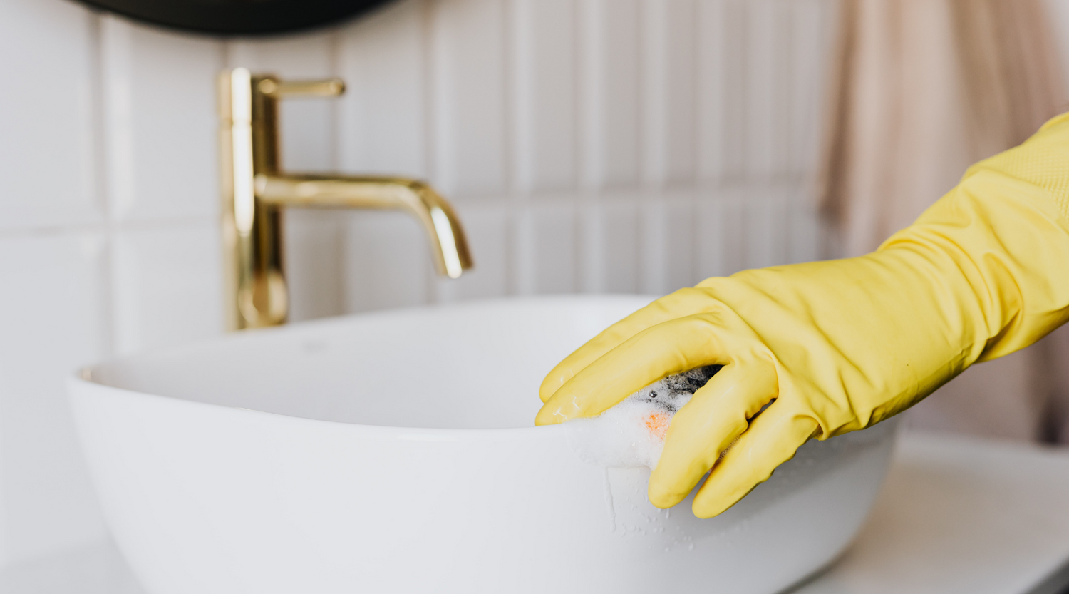 Hand wearing a yellow glove cleaning a white sink with a gold faucet.