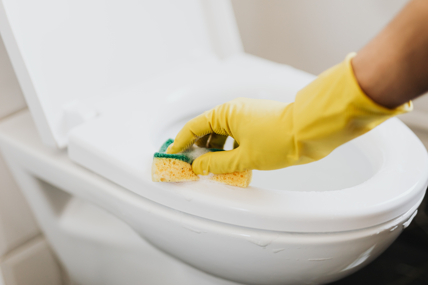 Person cleaning a toilet seat with a yellow glove and sponge