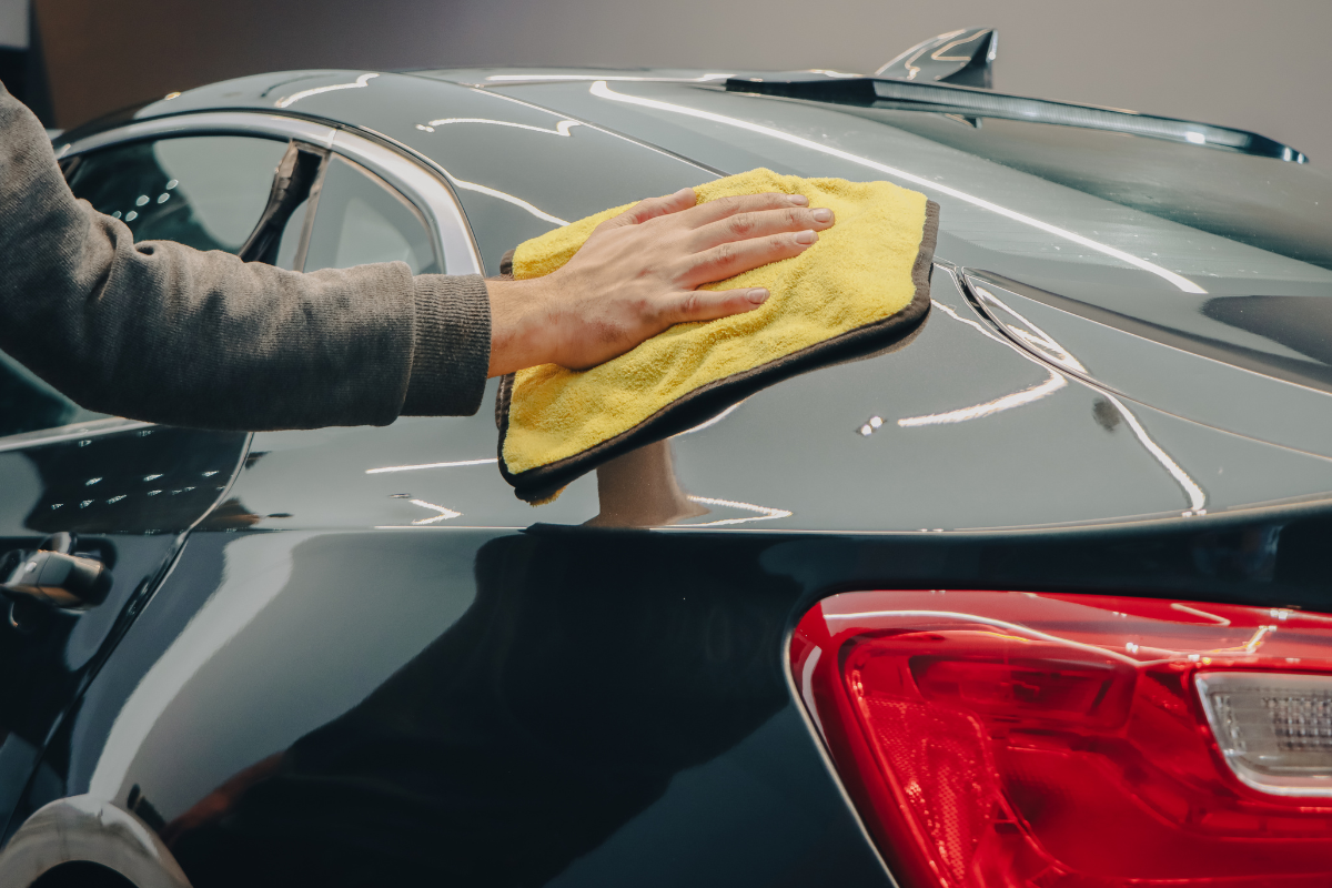 A close-up of a person's hand and arm wiping a glossy black car trunk with a yellow microfiber cloth in a garage setting.
