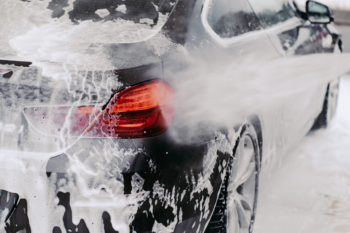 A close-up action shot of a black car covered in white foam, with steam or mist rising from a high-pressure washer nozzle directed at the rear quarter panel and taillight area in a snowy outdoor setting.