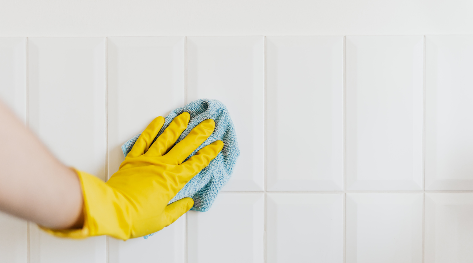 Hand wearing a yellow glove cleaning a white tiled floor with a blue cloth.
