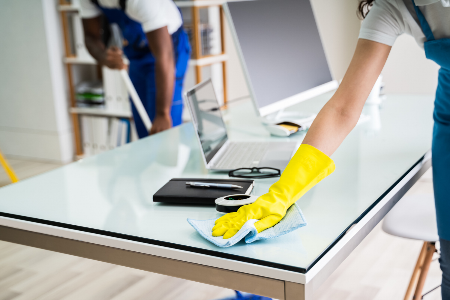 Person cleaning a glass desk with a yellow glove in an office setting