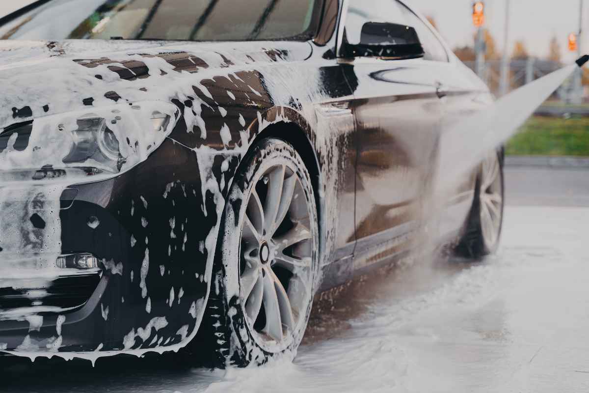 A black car covered in white soap foam, being rinsed with a high-pressure water spray on an outdoor street.