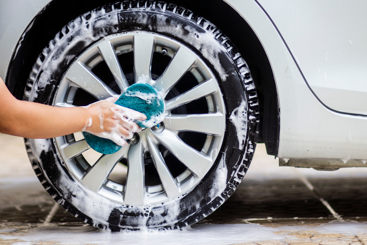 A close-up of a car wheel being washed with soapy foam while a person's hand scrubs the silver alloy rim with a blue sponge.